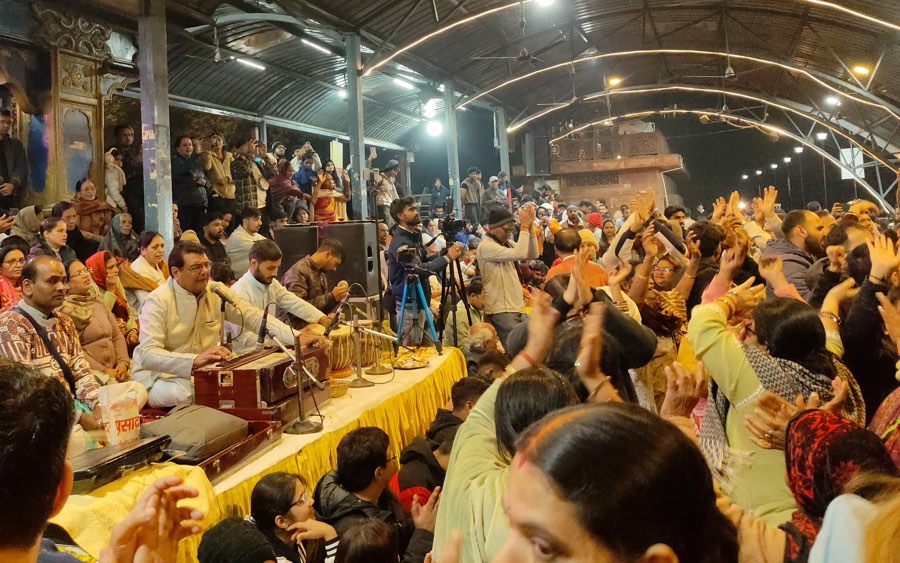 A devotional singing gathering: a singer at the harmonium with a tabla player, and a crowd with hands raised